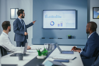 Business meeting with a presenter in a suit pointing at a large screen displaying charts and graphs.