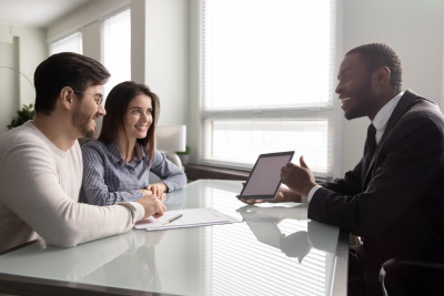 a couple consults with a professional in an office, reviewing documents and discussing information on a tablet.