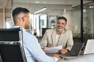 Two men in a modern office setting engaged in a discussion, with laptops and documents on the table.