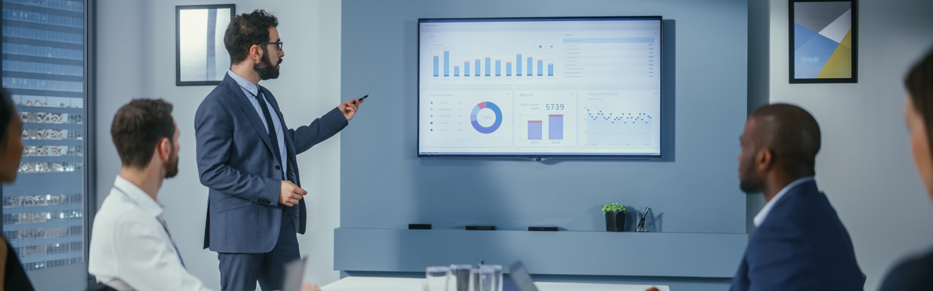 Business meeting with a presenter in a suit pointing at a large screen displaying charts and graphs.