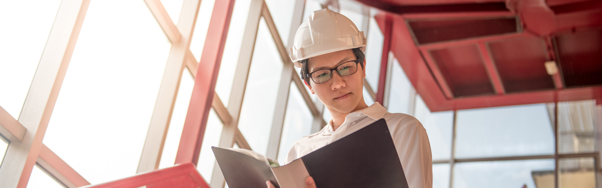 Construction worker wearing a white hard hat and shirt, reading a black folder in a sunlit building.