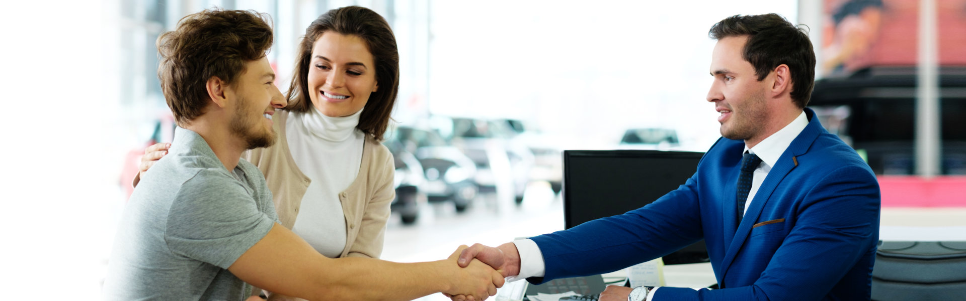 A couple shakes hands with a salesman in a blue suit at a car dealership, finalizing a purchase agreement.